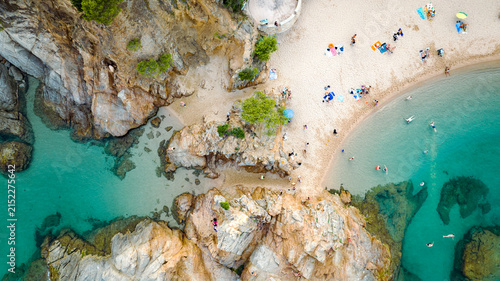 Aerial view of a sandy Mediterranean cove with rocky formations, clear turquoise water, swimmers and colorful beach umbrellas.
