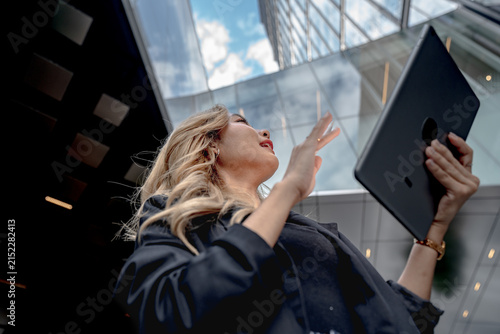 Foto Hair blowing in the wind, an Asian businesswoman raises her hand high beneath the glass atrium, tablet held aloft as she gazes upward energetically, on a modern city street