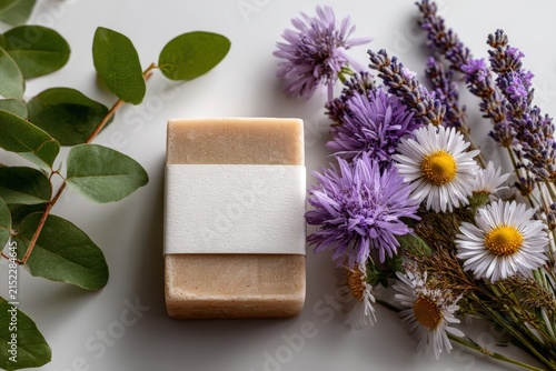 Natural handmade soap bar wrapped in white paper placed beside fresh lavender and daisies on a light surface with eucalyptus leaves for a calming aesthetic