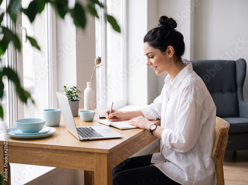 Young woman working remotely from home, writing in a notebook and using a laptop at a bright wooden table by a window.