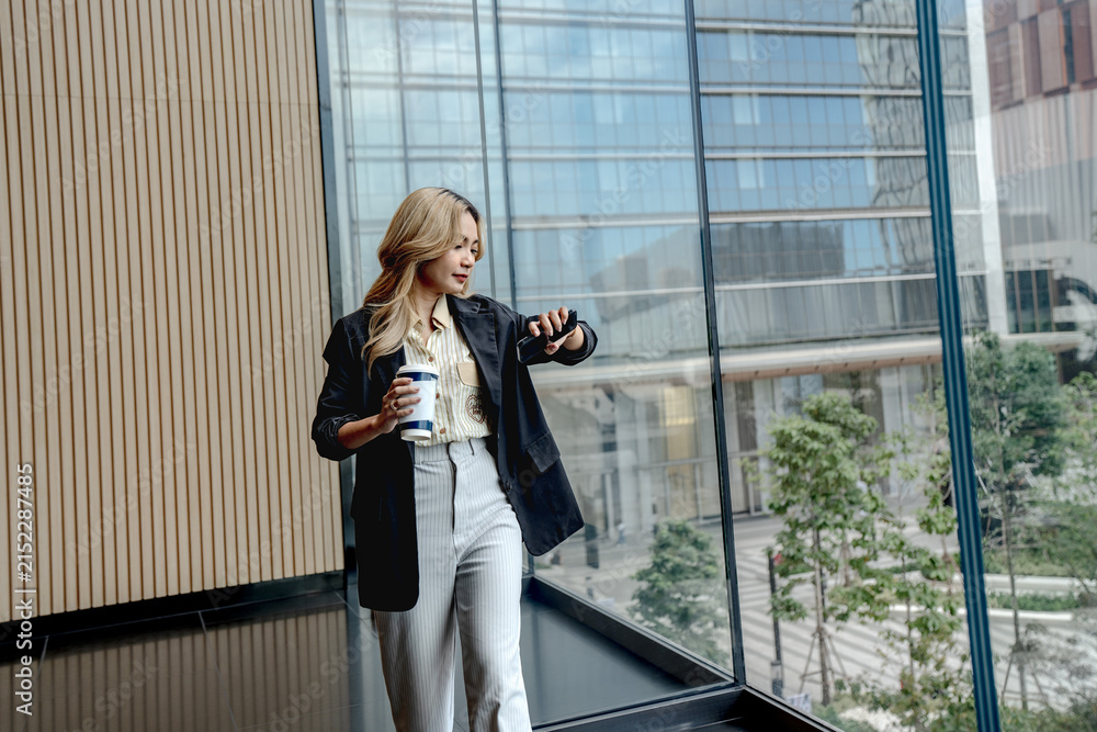 Fototapeta premium Businesswoman raises her smartphone to check the screen with a serious expression, coffee cup held low, leaning against the glass wall of a Bangkok office tower.