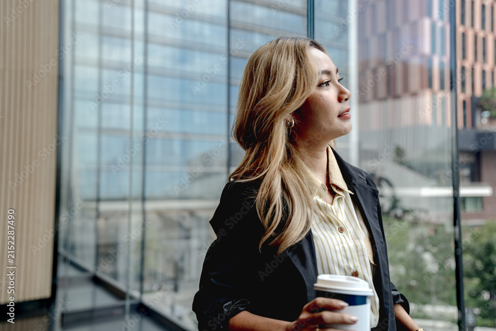 Fototapeta premium Tight close up portrait of a businesswoman in profile gazing serenely into the distance, coffee cup held low as soft light reflects off the Bangkok tower glass.