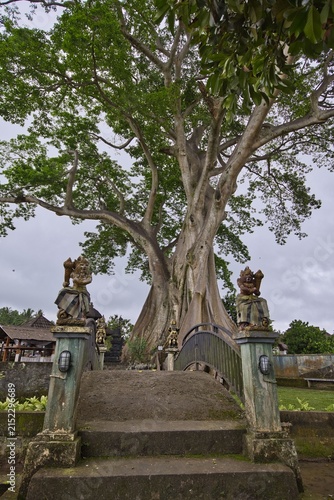 Foto A traditional stone bridge with guardian statues leads towards a magnificent, sacred giant banyan tree at the historic Pura Mengening temple complex in Bali, Indonesia