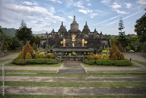 Fotografie Magnificent mini replica of Borobudur temple surrounded by lush gardens, pathways, and golden Buddha statues under a blue sky at Brahma Vihara Arama monastery in Bali