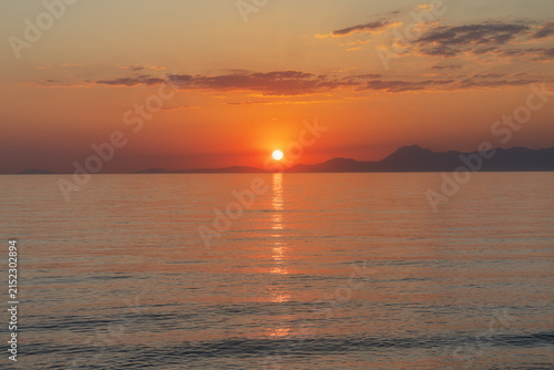 Bright orange sunset over calm sea with mountain silhouettes on horizon and sun path reflection on water.