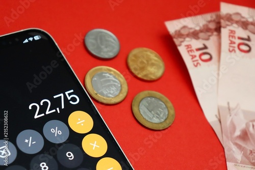 Smartphone displaying a calculator app next to Brazilian Real banknotes and coins. Brazilians financial planning and budgeting.