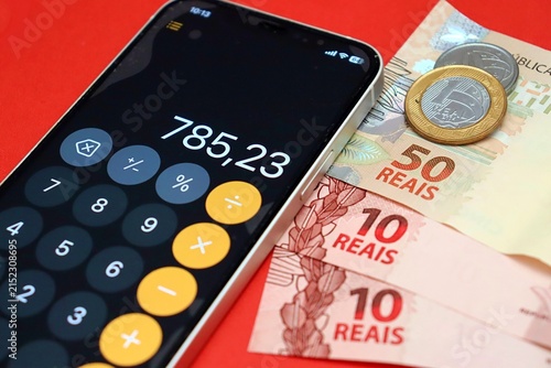 Smartphone displaying a calculator app next to Brazilian Real banknotes and coins. Brazilians financial planning and budgeting.