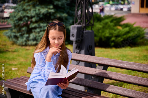 Принти на полотні A 12‑year‑old girl with light brown hair is sitting on a bench, engrossed in reading a book