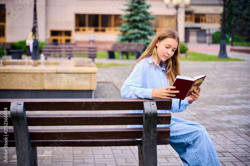 Fotografie A 12‑year‑old girl with light brown hair is sitting on a bench, engrossed in reading a book