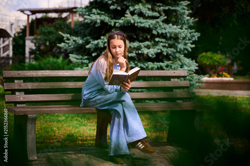 Obraz na płótnie A 12‑year‑old girl with light brown hair is sitting on a bench, engrossed in reading a book