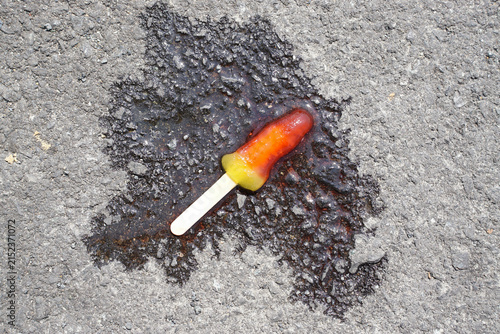 Melted colorful popsicle on a wooden stick on rough asphalt under bright sunlight
