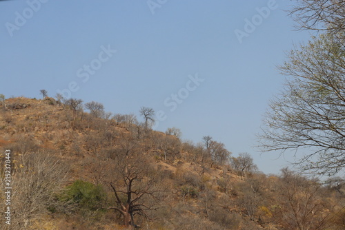 Scenic semi-arid mountainous landscape featuring rugged rocky hills, dry woodland, scattered trees, large natural boulders and a tranquil reservoir surrounded by rolling terrain. The collection captur