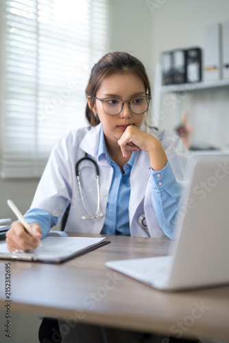 Young Asian female doctor writing medical notes while reviewing patient records on laptop in bright clinic office