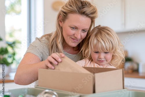 Young Caucasian mother and child sorting household waste in a kitchen, with a cardboard box and various recyclable materials visible on the table