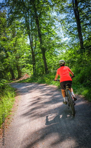 female Cyclist riding a bicycle along a peaceful forest avenue surrounded by lush green trees and dappled sunlight. Stuttgart, Baden-Württemberg, Germany.