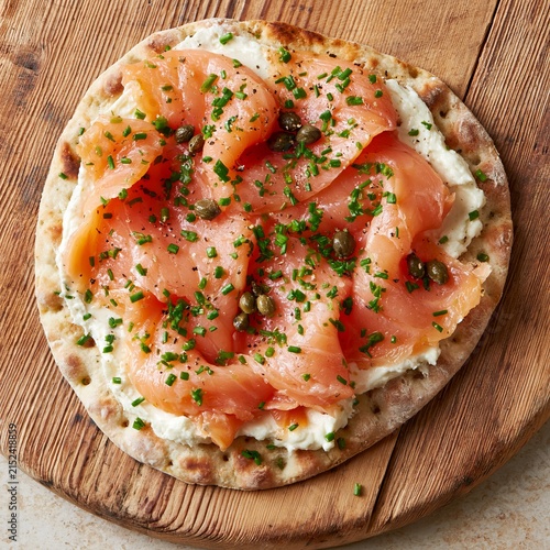 Overhead shot of salmon and cream cheese flatbread with capers and chives on a wooden board, showcasing a delicious culinary presentation