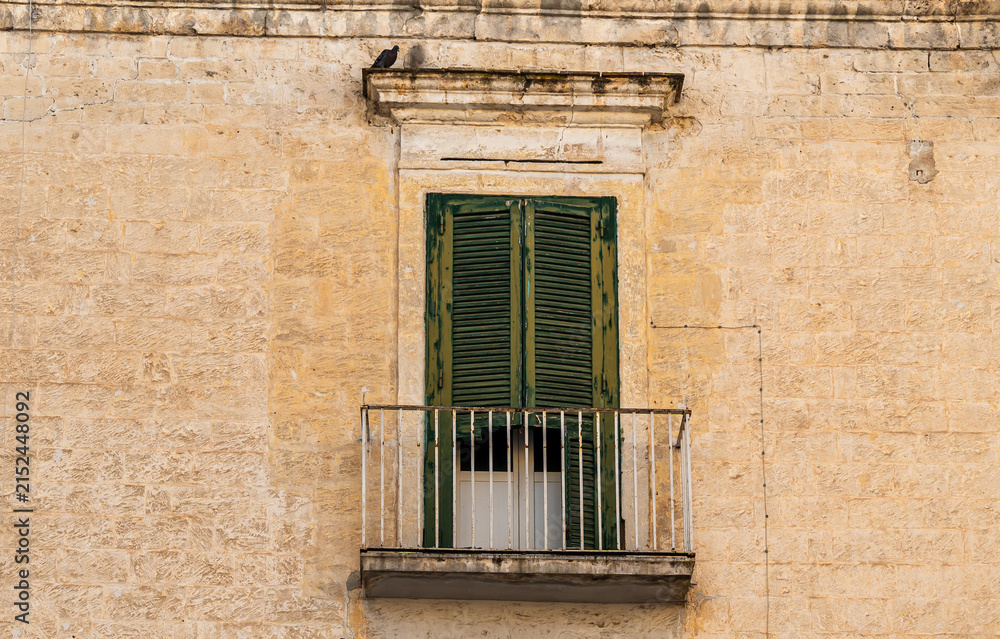 Fototapeta premium Old stone building facade in Bari, Italy with weathered green wooden shutters on balcony door, rustic limestone wall texture and iron railing, nobody