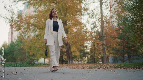 Barefoot young woman in light pantsuit moves along leaf-covered park path, carrying high heels in one hand and showing calm transition from formal to relaxed style in warm daylight