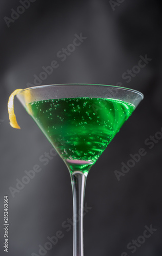 Spooky green martini cocktail decorated with lemon peel, surrounded by subtle white smoke fog on a dark, moody background, perfect for Halloween bar menus, witchy potion concepts