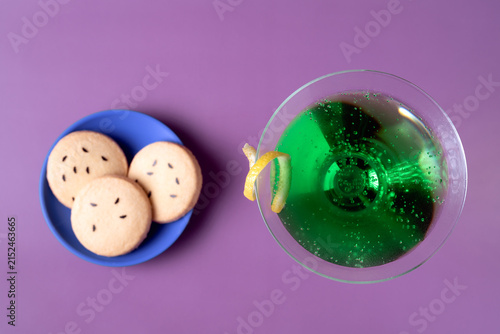 overhead flat lay of a green Halloween martini cocktail garnished with lemon peel alongside bat cookies set against a purple background with clean copy space for party invitations