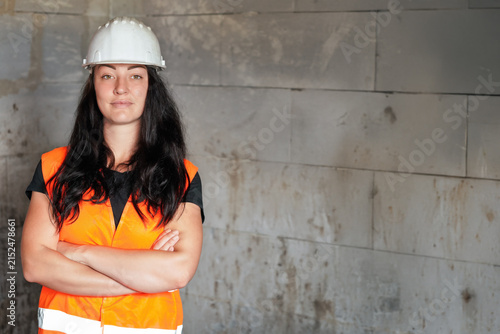 Young woman in builders hi vis vest and helmet posing next to bare bricks wall - females in construction concept