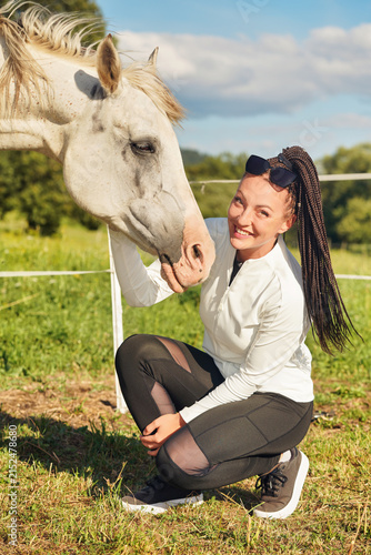 Young woman with afro braids style hair crouching next to her Arabian white horse, outside photo in bright daylight sun