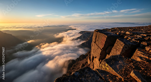 Mountain cliff edge above clouds during sunrise with golden light and vast landscape