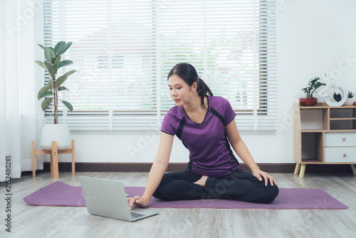 Foto Online Yoga Session: A woman with Asian ethnicity sits in a meditative pose on a yoga mat, engaging with her laptop, suggesting an online yoga or fitness class