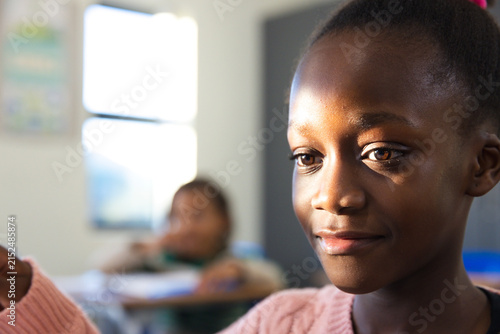 School-aged female child sitting at desk in classroom wearing pink sweater holding notebook