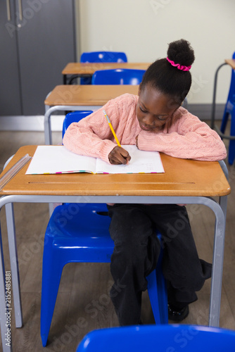 African American child in pink sweater writing in notebook at desk in classroom with yellow pencil