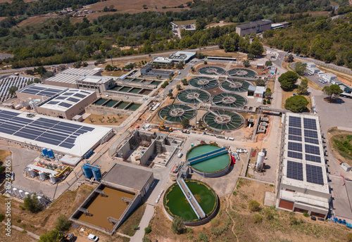 Top-down view of a drinking water treatment facility featuring circular flocculation clarifiers, rectangular filtration basins, blue storage tanks, and rooftop solar panels surrounded by countryside