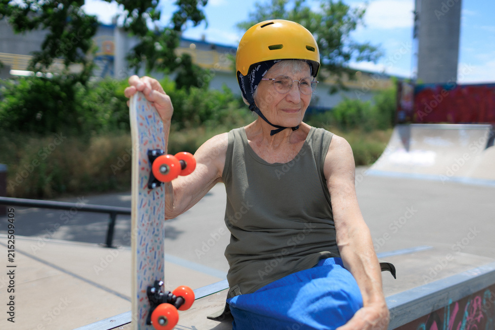 Fototapeta premium Elderly woman wearing a helmet is holding her skateboard in skate park