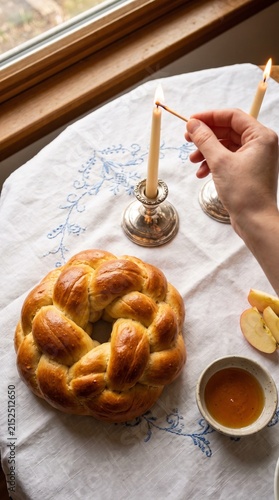 Hand lighting Jewish holiday candles beside round challah apples and honey on a festive table during Rosh Hashanah