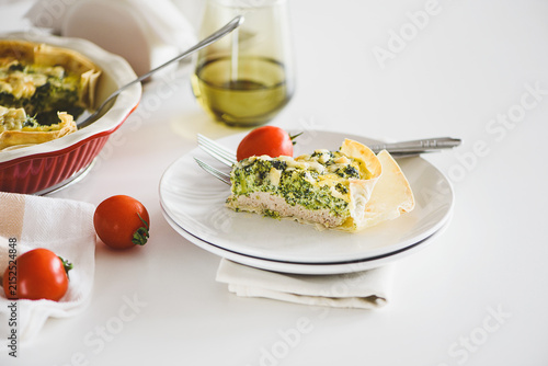 Freshly baked broccoli quiche slice with cherry tomatoes served on a white plate with a fork and glass of olive oil