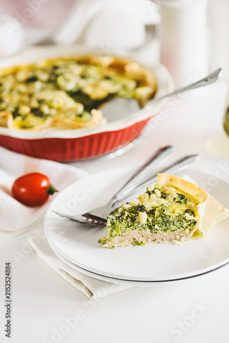 Freshly Baked Broccoli Quiche Slice on a Plate with Whole Quiche Background and Tomato