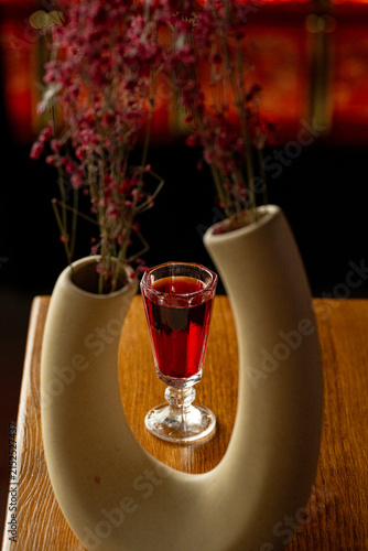 A glass filled with a red drink sits on a wooden table. A curved vase holds flowers, creating a simple arrangement in a cafe. The background is softly lit