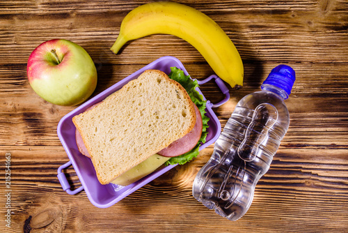Ripe apple, banana, bottle of water and lunch box with sandwiches on a wooden table. Top view