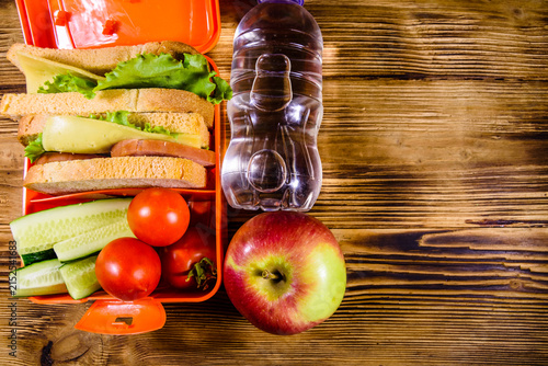 Ripe apple, bottle of water and lunch box with sandwiches, cucumbers and tomatoes on wooden table. Top view