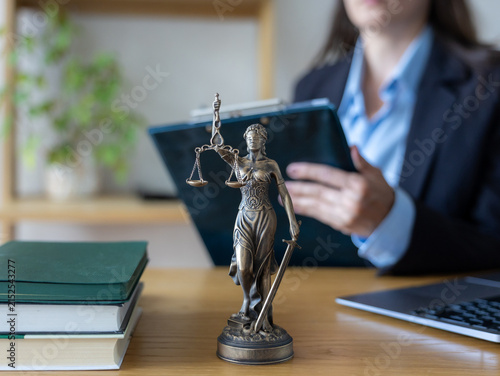 Woman lawyer in glasses using blue smartphone at desk. Bronze Justice statue, green books near open laptop. Focused legal professional handling online client consultations using modern digital tools.