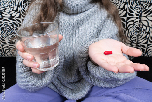 Quadro su tela Child in warm knitted sweater holding a red capsule pill in open palm and a glas
