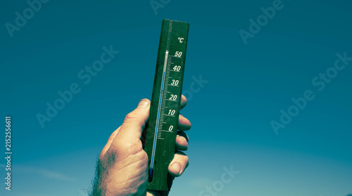 Hand holding wooden outdoor thermometer against clear blue sky showing summer heat