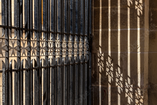 Wrought-iron railings with Maltese cross motifs cast dramatic patterned shadows on a limestone wall in Malta