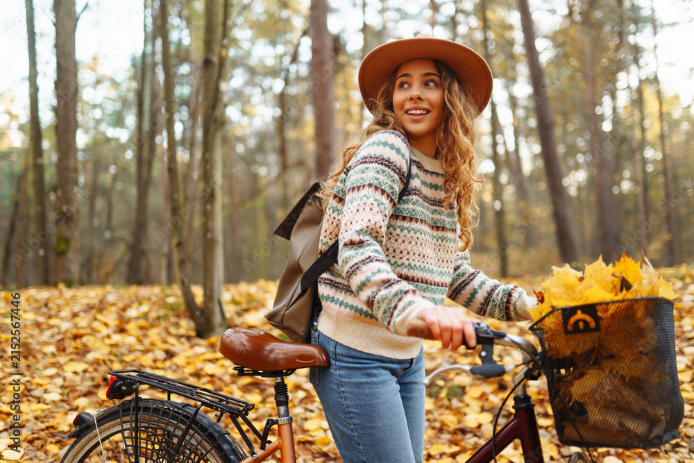 Fototapeta premium Young woman wearing hat and carrying backpack rides bicycle in autumn forest. Woman with bicycle in clearing surrounded by colorful fallen leaves outdoors. Concept of weekends, relaxation.