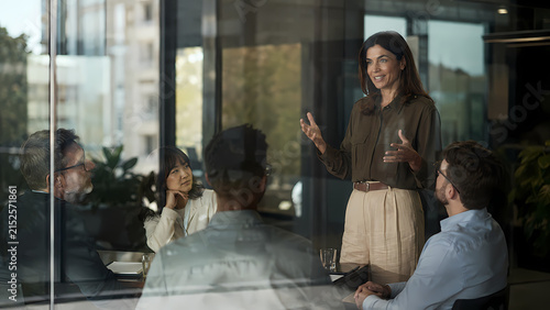 Happy middle aged business woman executive leader presenting project management planning and corporate strategy to diverse colleagues during professional team meeting in modern glass office boardroom