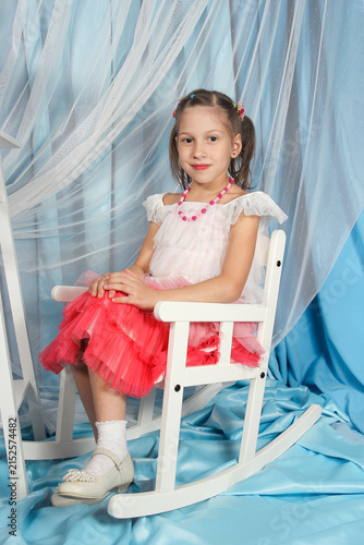 Charming Little Girl in White Dress Relaxing on Vintage Rocking Chair with a Shy Smile