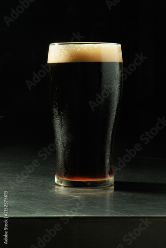 Fotografía Full pint of dark craft beer served in a chilled glass with a creamy foam head against a black background