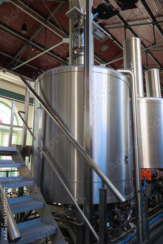 Large stainless steel brewing tanks and industrial fermentation equipment inside a modern brewery production facility. Vertical view of polished metal vessels, pipes, and access stairs in a clean