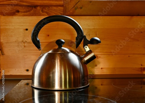 A shiny stainless steel whistling tea kettle sits on a modern induction stovetop against a warm rustic wooden wall background, ready for boiling water and making hot beverages.