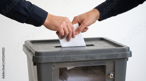 A close-up of two hands, one placing a ballot into a grey ballot box. The image depicts the act of voting and democratic participation