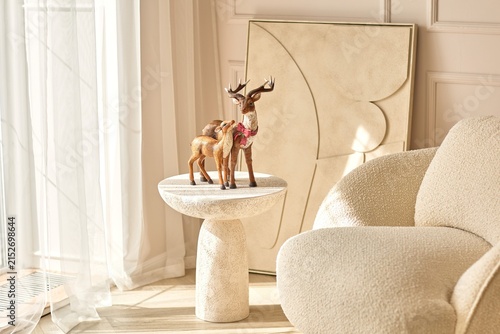 Decorative deer figurines stand on a side table beside a cream armchair in a sunlit livingroom interior with neutral decor, textured furniture, abstract artwork and soft curtains.